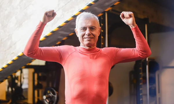 Homem idoso em uma academia, vestindo uma roupa esportiva vermelha, com as duas mãos levantadas em forma de comemoração, ao lado de equipamentos e luzes ao fundo.