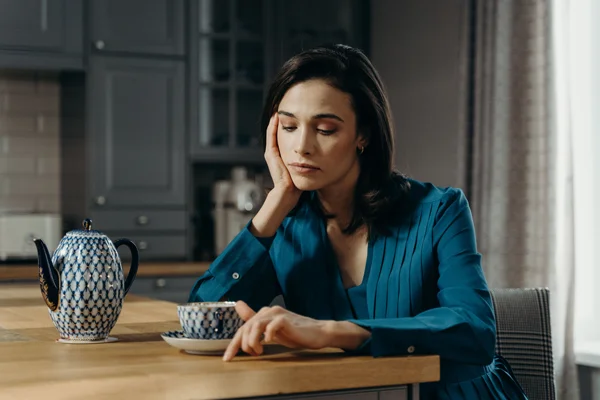 Mulher em ambiente doméstico, sentada à mesa, com xícara e bule estilizado ao lado, apoiando o rosto com a mão e aparentando cansaço ou preocupação.