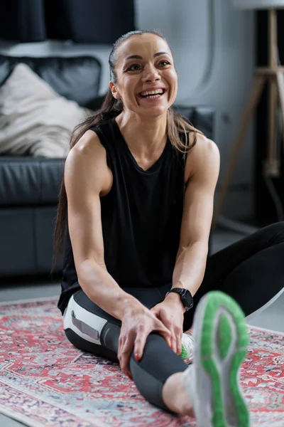 Mulher sorridente alongando em casa, sentada no chão com uma perna estendida, usando roupa esportiva escura e tênis com solado verde, em ambiente moderno e aconchegante.
