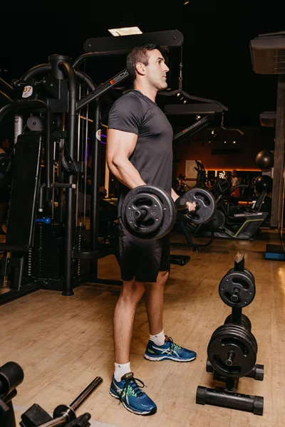 Homem em uma academia realiza levantamento de peso com halteres, em pé ao lado de equipamentos de musculação, com discos de peso no chão e iluminação de estúdio