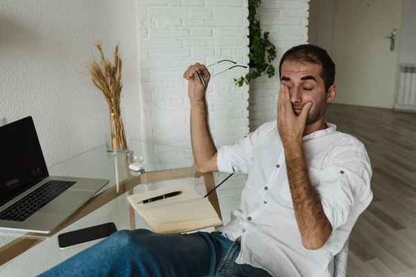 Homem sentado em uma sala de home office, segurando o rosto e demonstrando cansaço enquanto trabalha em uma mesa com notebook, documentos e copo de vidro, com planta decorativa ao fundo.